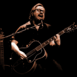 Artisti Heikki Hela tummasävyisessä kuvassa kitaran kanssa