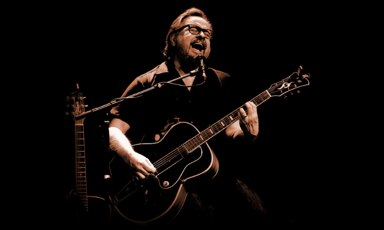 Artisti Heikki Hela tummasävyisessä kuvassa kitaran kanssa