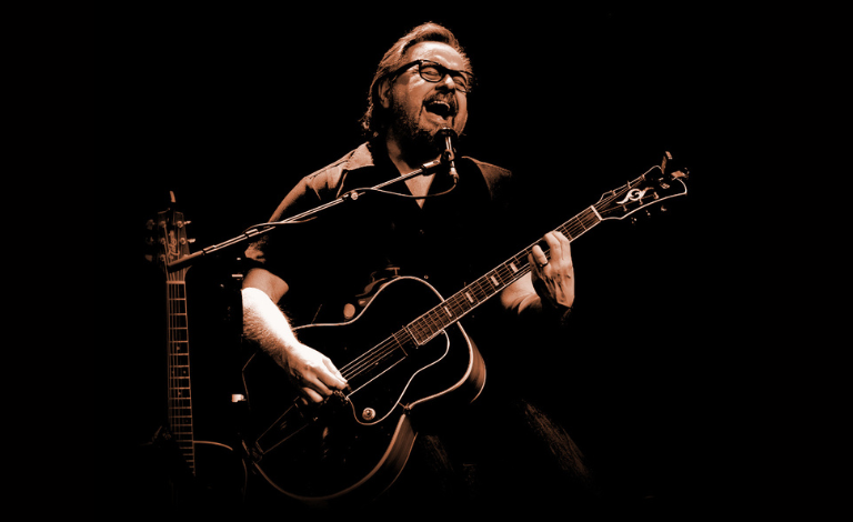 Artisti Heikki Hela tummasävyisessä kuvassa kitaran kanssa